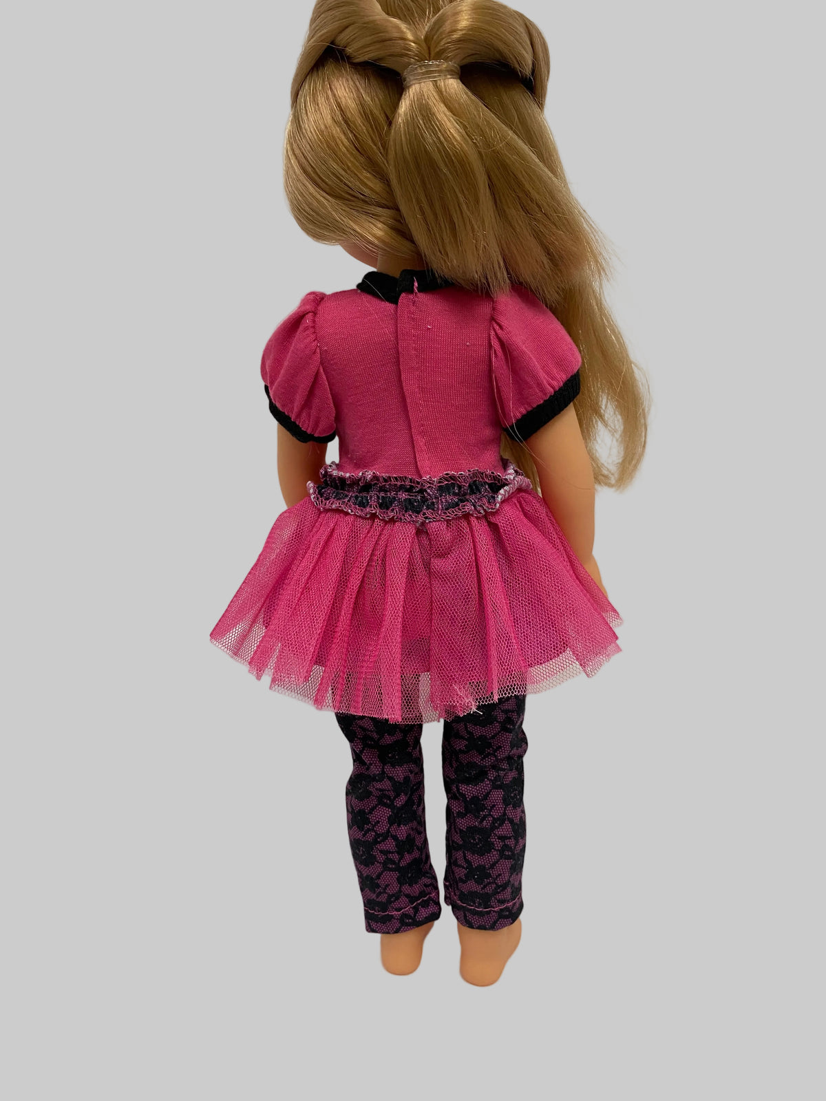 A doll viewed from the back wearing a bright pink short-sleeve top with black trim, a pink tutu skirt, and dark patterned leggings, against a plain light gray background.